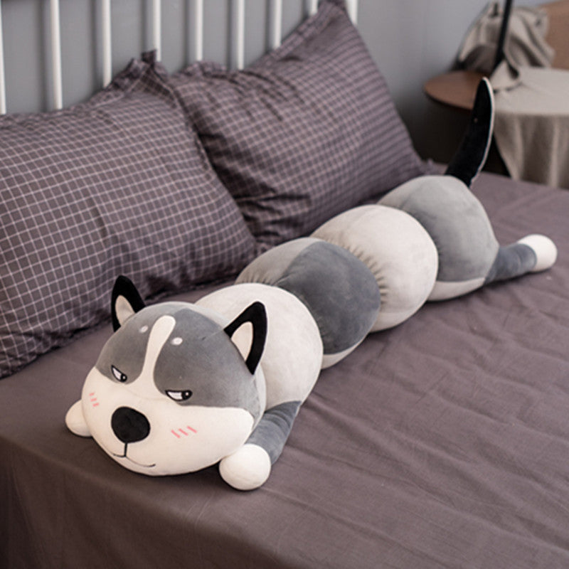 Long plush toy shaped like a dog lying on a bed with pillows in the background.